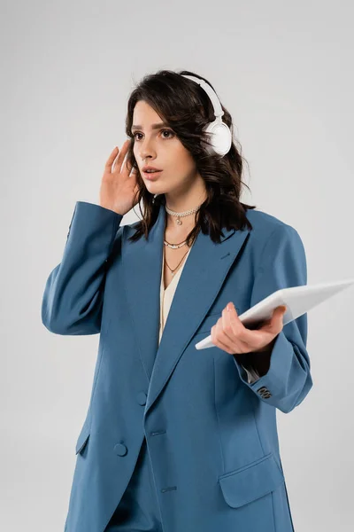 Tense woman in blue blazer adjusting wireless headphones while holding digital tablet isolated on grey — Stock Photo