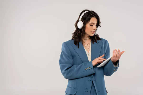 Mujer morena en auriculares inalámbricos y blazer azul usando tableta digital aislada en gris - foto de stock