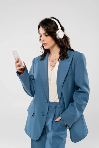 Trendy woman in wireless headphones standing with hand in pocket of blue pants and looking at smartphone isolated on grey — Stock Photo