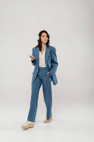 Full length of woman in wireless headphones and blue suit walking with smartphone and hand in pocket on grey — Stock Photo