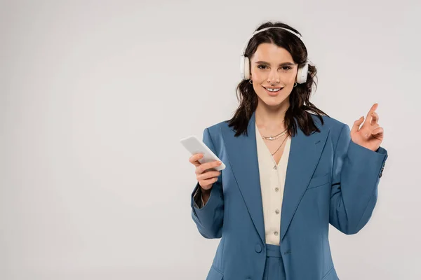 Pleased brunette woman in wireless headphones holding smartphone and pointing with finger isolated on grey — Stock Photo