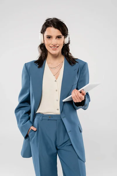 Happy woman in blue suit and wireless headphones standing with hand in pocket and digital tablet isolated on grey — Stock Photo