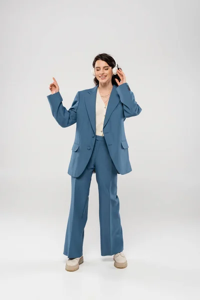 Longitud completa de mujer alegre en traje azul escuchando música en auriculares inalámbricos sobre fondo gris — Stock Photo