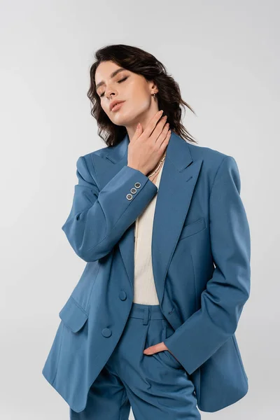Sensual brunette woman in blue suit holding hand in pocket and touching neck while standing with closed eyes isolated on grey — Stock Photo