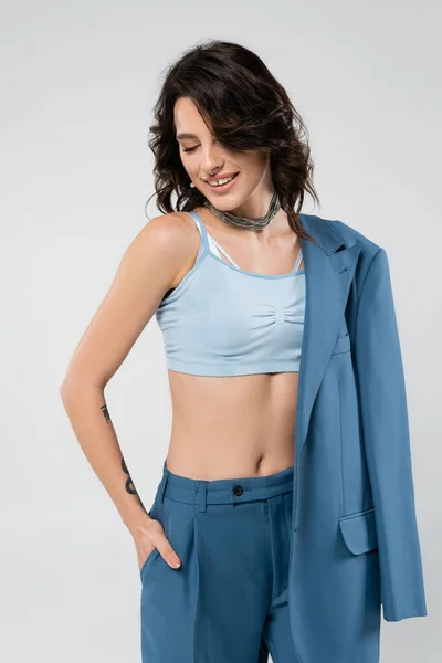 Smiling woman in blue crop top standing with hand in pocket of trousers isolated on grey — Stock Photo