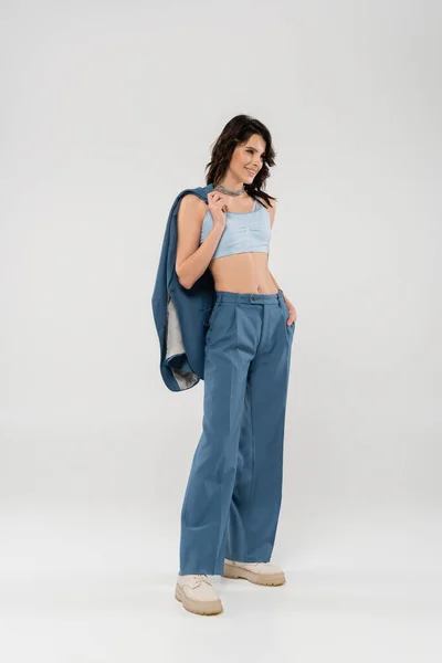 Full length of trendy woman in blue pants and crop top standing with jacket on grey background — Stock Photo
