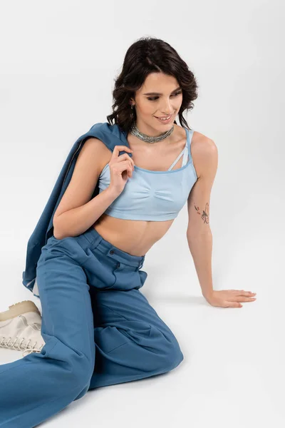 Happy woman in crop top and blue trousers holding jacket while sitting on grey — Stock Photo