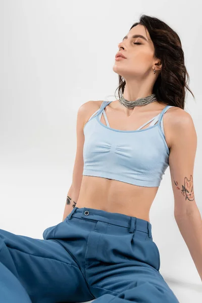 Tattooed woman in blue crop top and trousers sitting with closed eyes on grey background — Stock Photo