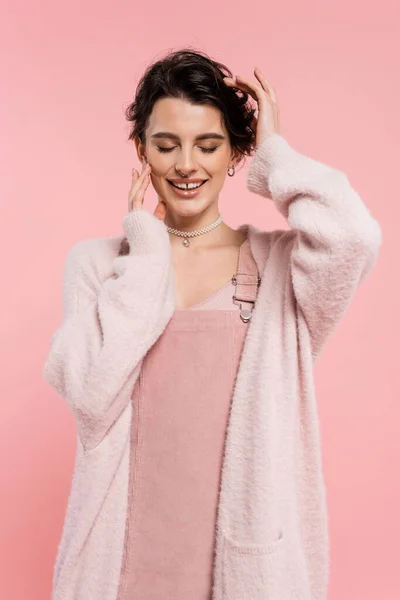 Happy brunette woman in warm cardigan and strap dress touching hair and face while posing with closed eyes isolated on pink — Stock Photo