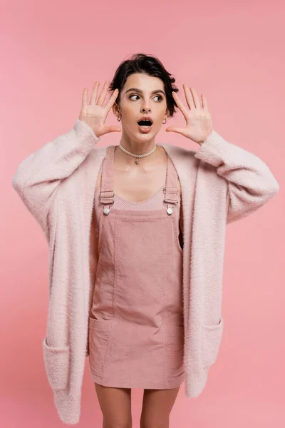 Shocked woman in strap dress and warm cardigan standing with open mouth and showing wow gesture isolated on pink — Stock Photo