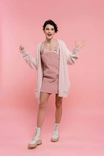 Full length of excited woman in strap dress and fluffy cardigan showing wow gesture and looking away on pink background — Stock Photo
