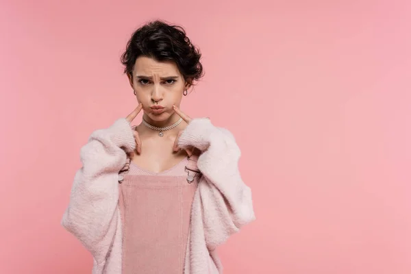 Offended woman in fluffy cardigan touching puffed cheeks while looking at camera isolated on pink — Stock Photo