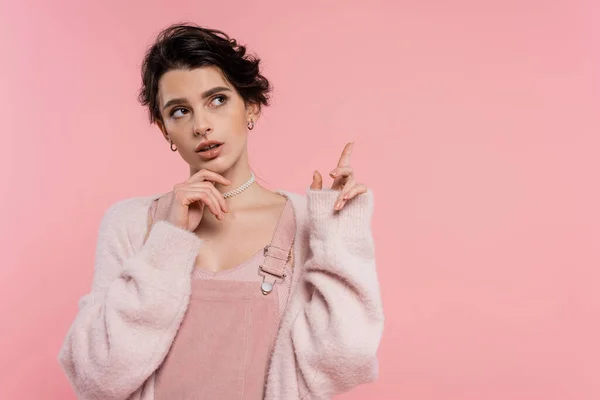 Thoughtful brunette woman touching chin and pointing with finger isolated on pink — Stock Photo