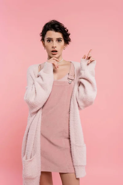 Astonished woman in cozy cardigan touching chin and pointing with finger isolated on pink — Stock Photo