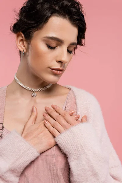 Jeune femme en collier de perles et piercing posant avec les mains sur la poitrine et les yeux fermés isolés sur rose — Photo de stock