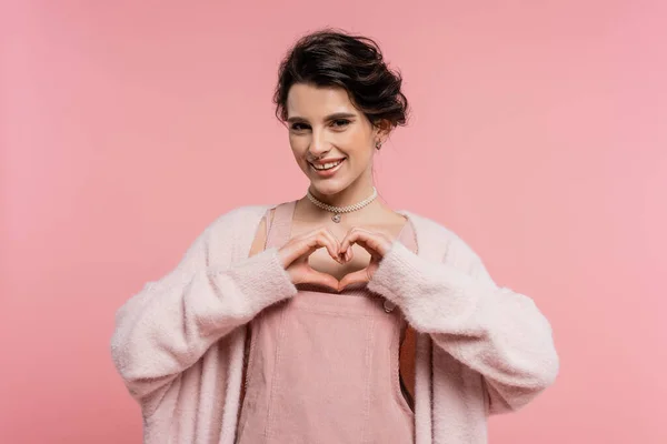 Donna felice in cardigan caldo e soffice che mostra il segno del cuore con le mani isolate sul rosa — Foto stock