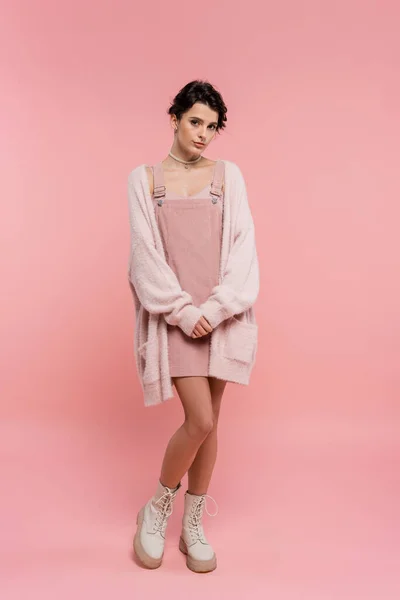 Volle Länge der brünetten Frau in Stiefeln und Trägerkleid mit warmer Strickjacke Blick in die Kamera auf rosa Hintergrund — Stockfoto