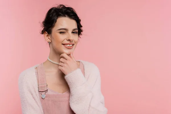Tricky and cheerful woman in fluffy cardigan touching chin and winking at camera isolated on pink — Stock Photo