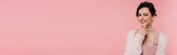 Knifflige und lächelnde Frau berührt Kinn und zwinkert Kamera isoliert auf rosa, Banner — Stockfoto