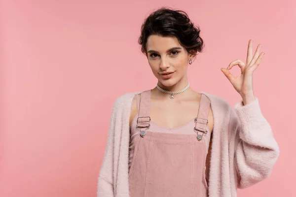 Smiling brunette woman in strap dress and fluffy cardigan showing okay sign isolated on pink — Stock Photo