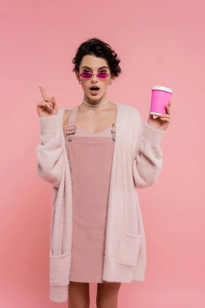 Femme étonnée dans un cardigan chaud et des lunettes de soleil tenant tasse en papier et pointant avec le doigt isolé sur rose — Photo de stock