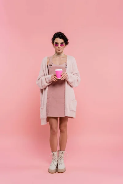 Full length of woman in warm cardigan and trendy sunglasses holding coffee to go on pink — Stock Photo