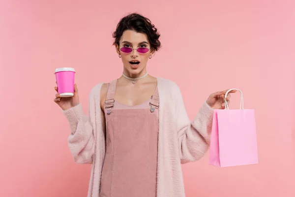 Femme étonnée dans des lunettes de soleil et cardigan chaud tenant sac à provisions et tasse en papier isolé sur rose — Photo de stock