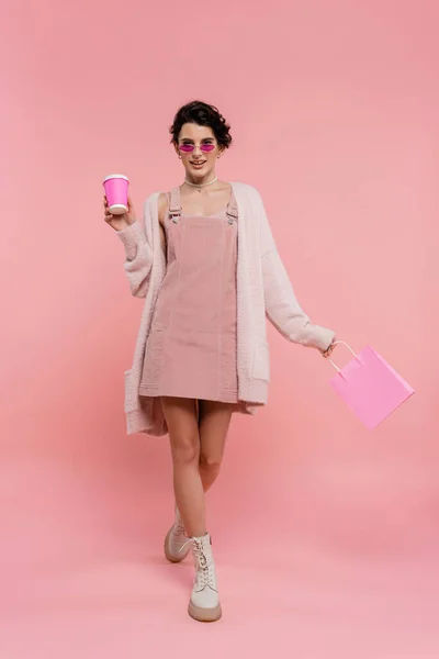 Pleine longueur de femme à la mode marchant avec tasse en papier et sac à provisions sur rose — Photo de stock