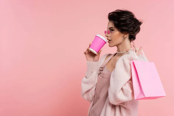 Mulher morena na moda óculos de sol beber café para ir enquanto segurando saco de compras isolado em rosa — Fotografia de Stock