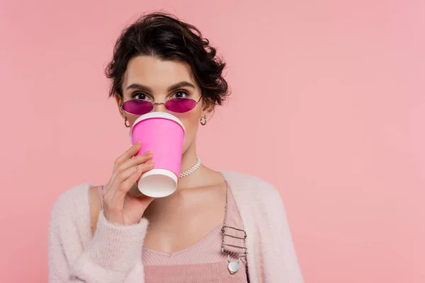 Brunette woman in trendy sunglasses drinking coffee from paper cup isolated on pink — Stock Photo