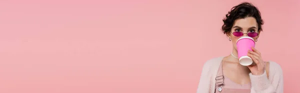 Young woman in stylish sunglasses drinking coffee and looking at camera isolated on pink, banner — Stock Photo