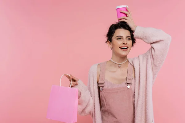 Femme joyeuse et élégante avec sac à provisions tenant tasse en papier sur la tête isolé sur rose — Photo de stock