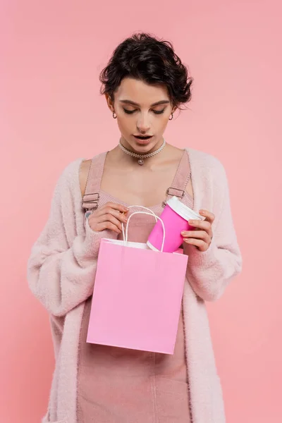 Morena mujer en acogedor cárdigan poner taza de papel en bolsa aislada en rosa - foto de stock