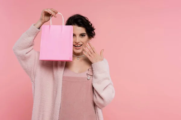 Femme heureuse dans un cardigan doux montrant sac à provisions et tenant la main près du visage isolé sur rose — Stock Photo