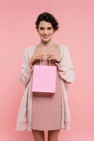 Pleased brunette woman in warm cardigan holding shopping bag and smiling at camera isolated on pink — Stock Photo