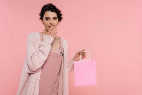 Femme étonnée dans un cardigan chaud tenant un sac à provisions et couvrant la bouche ouverte avec la main isolée sur rose — Photo de stock