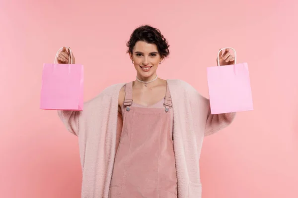 Smiling woman in trendy strap dress and fluffy cardigan showing shopping bags isolated on pink — Stock Photo