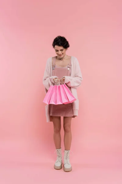 Pleine longueur de femme heureuse en cardigan chaud et bottes regardant les sacs à provisions sur fond rose — Photo de stock