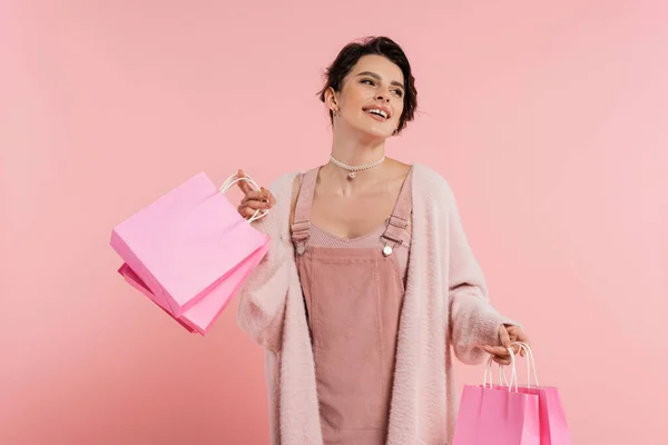 Mujer excitada en vestido de correa y acogedor cárdigan sosteniendo bolsas de compras y mirando hacia otro lado aislado en rosa - foto de stock