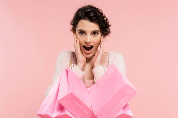 Astonished woman with shopping bags touching face and looking at camera isolated on pink — Stock Photo