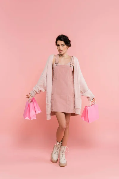 Pleine longueur de femme à la mode et surprise regardant la caméra tout en tenant des sacs à provisions sur rose — Photo de stock