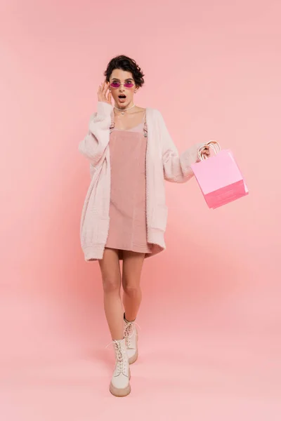 Femme à la mode et surprise touchant des lunettes de soleil tout en marchant avec des sacs à provisions sur rose — Photo de stock