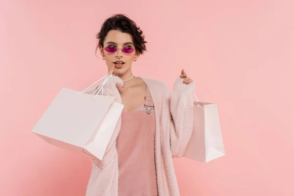 Verträumte Frau mit trendiger Sonnenbrille, Einkaufstüten in der Hand und isoliert auf rosa — Stockfoto