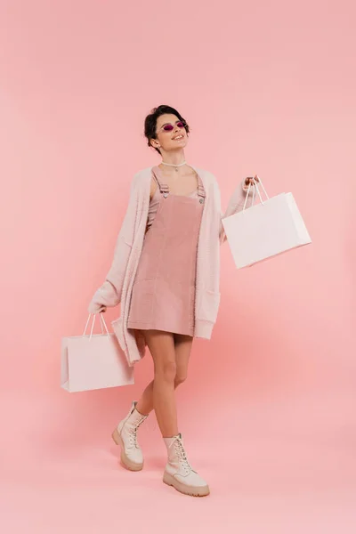 Lächelnde Frau mit Sonnenbrille und trendiger Kleidung, die mit Einkaufstaschen auf rosa — Stockfoto