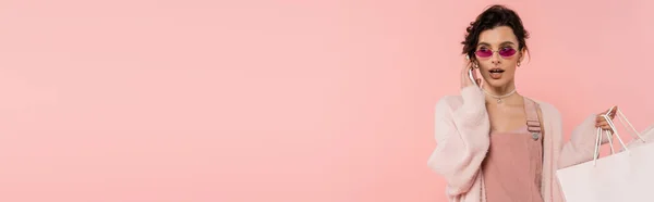 Mulher morena em casaco quente e óculos de sol segurando sacos de compras e falando no smartphone isolado em rosa, banner — Fotografia de Stock