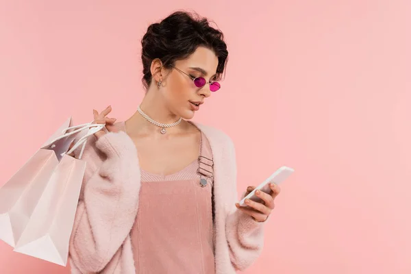 Mulher na moda óculos de sol segurando sacos de compras e mensagens no smartphone isolado em rosa — Fotografia de Stock