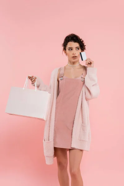 Donna bruna premurosa in cardigan caldo con borsa della spesa e carta di credito mentre distoglie lo sguardo isolato sul rosa — Foto stock