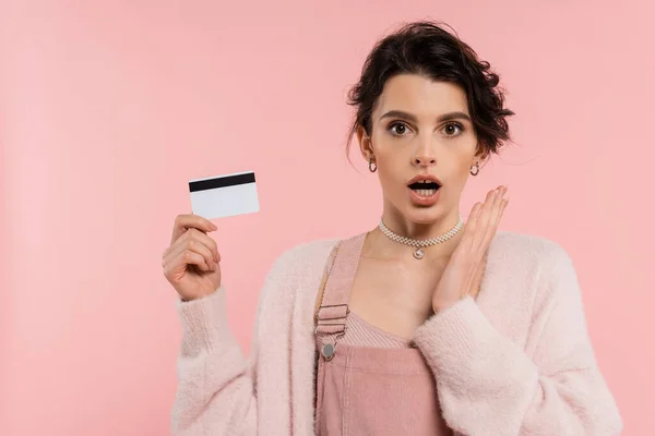 Femme étonnée tenant la main près du visage tout en montrant la carte de crédit isolé sur rose — Photo de stock