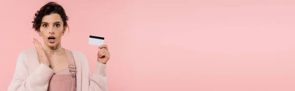 Femme étonnée dans le cardigan chaud tenant la carte de crédit et regardant la caméra isolée sur rose, bannière — Photo de stock