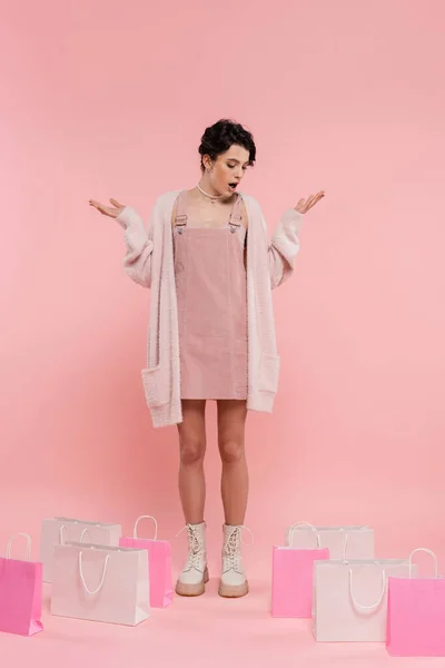 Longitud completa de la mujer sorprendida mostrando gesto wow mientras mira las bolsas de compras sobre fondo rosa - foto de stock